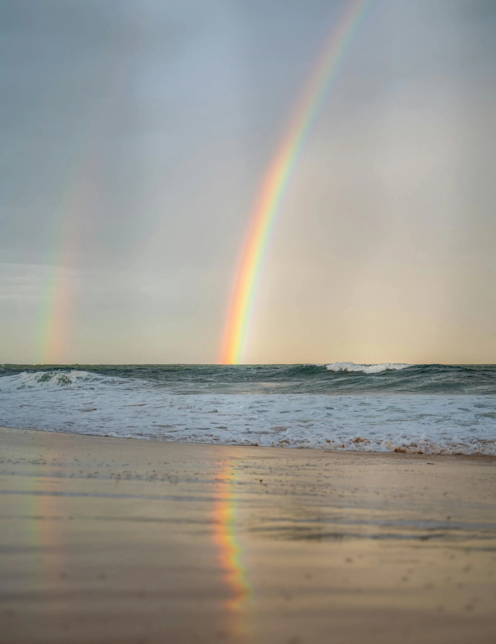 A beach scene with waves and wet sand, featuring a double rainbow in the sky.