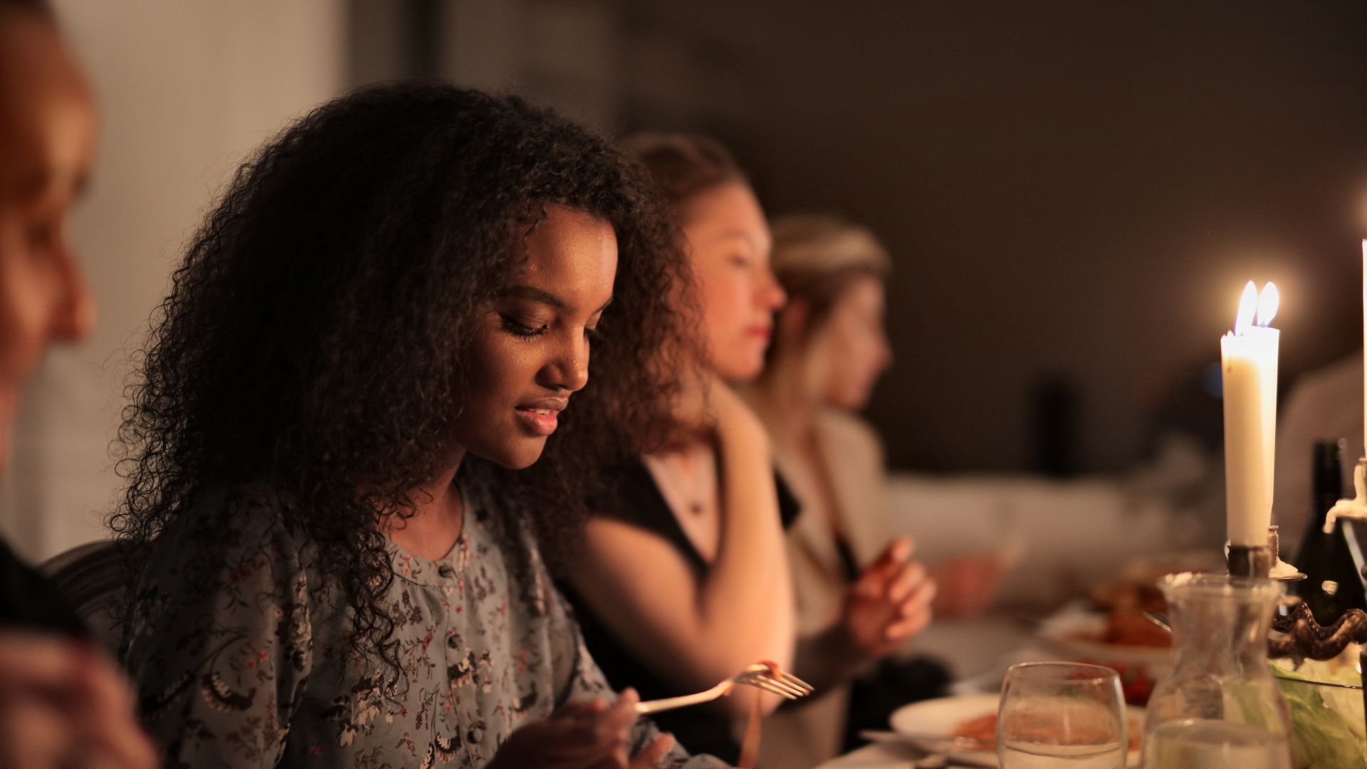Women sitting at a dinner table with lit candles, engaging in prayer before a meal, in a dimly lit room.