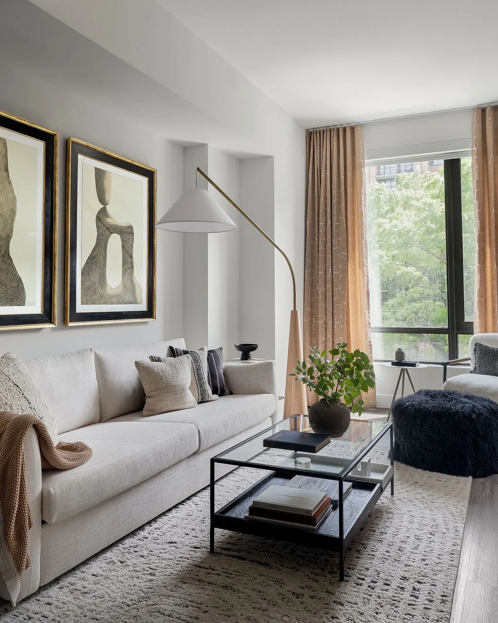 Living room with a white sofa, abstract art on the wall, a glass-top coffee table, a potted plant, large window with beige curtains, and a standing lamp.