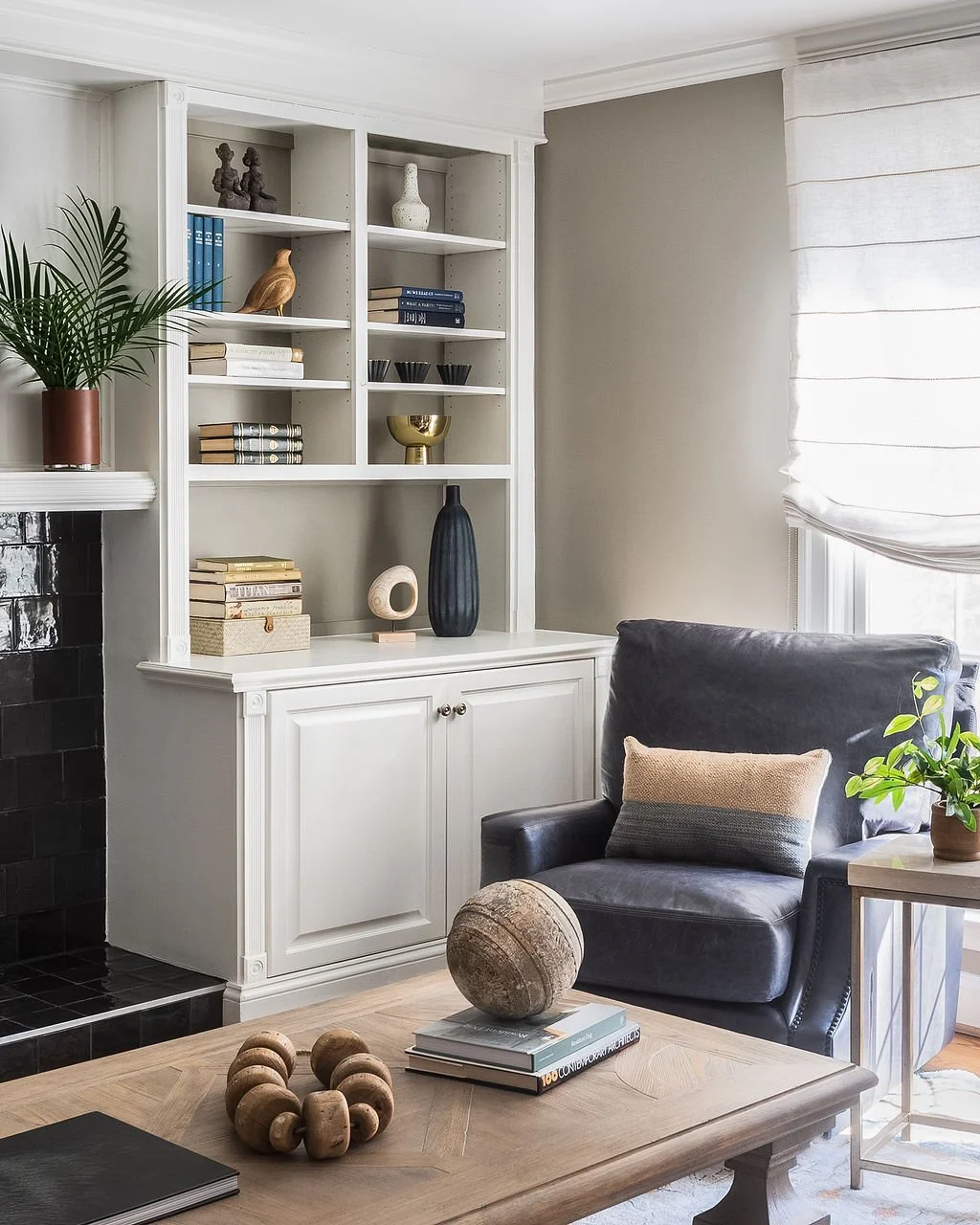 A cozy living room corner featuring a gray armchair with a beige pillow, a wooden coffee table with decorative objects, a white built-in bookshelf with decor, a small side table with a potted plant, and a window with white curtains.