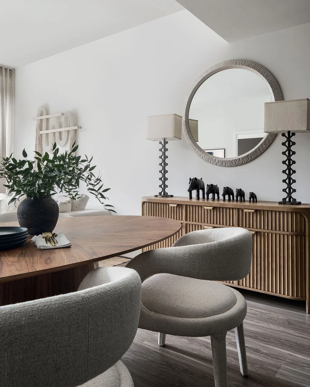 A dining room with a round wooden table, beige upholstered chairs, a large plant in a black vase, decorative black horse figurines, a wooden sideboard, two table lamps, and a round mirror on the wall.