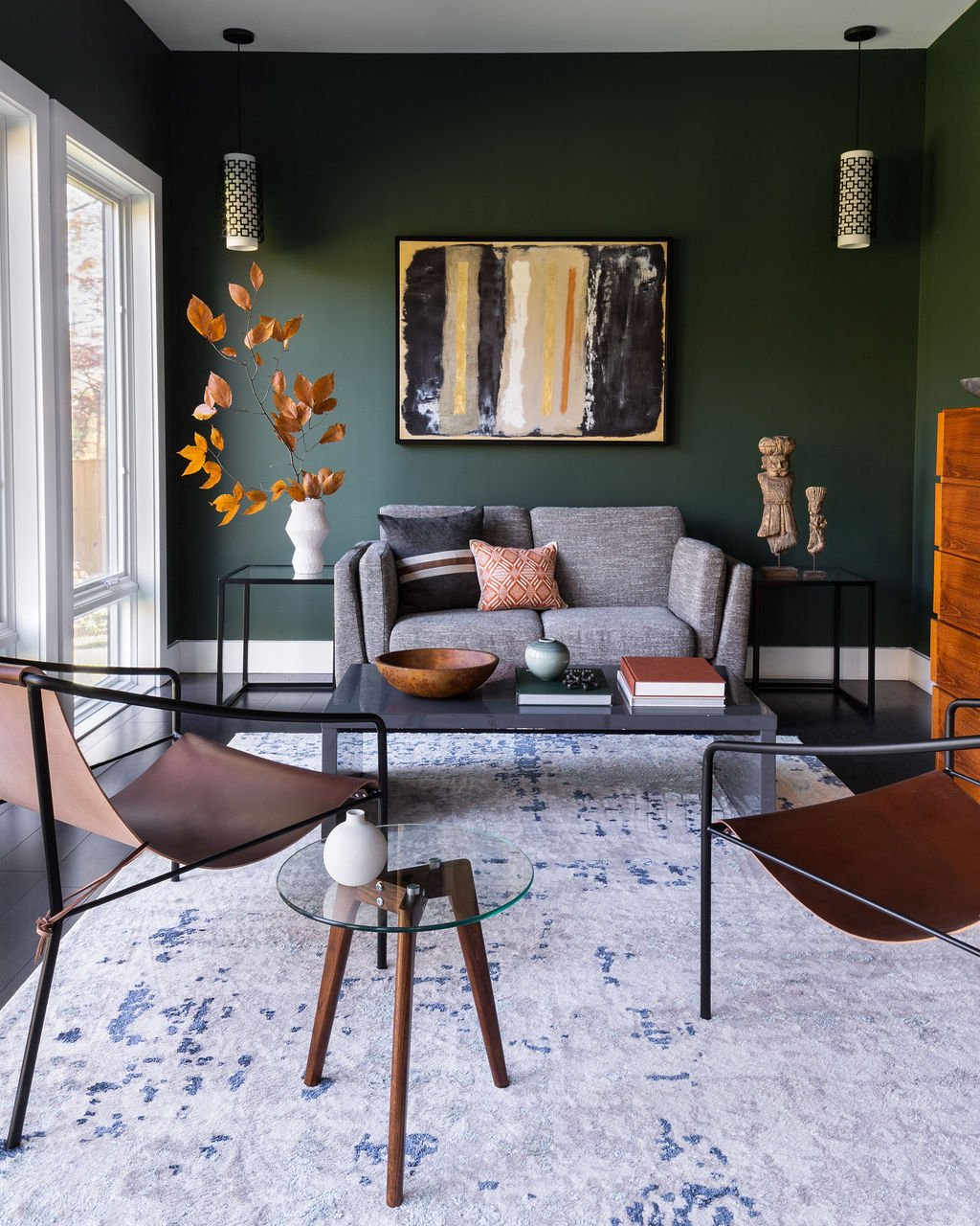 A modern living room with a dark green wall, framed abstract art, gray sofa, black coffee table with books and decorative items, two leather and metal lounge chairs, a large area rug, and string lights hanging from the ceiling.