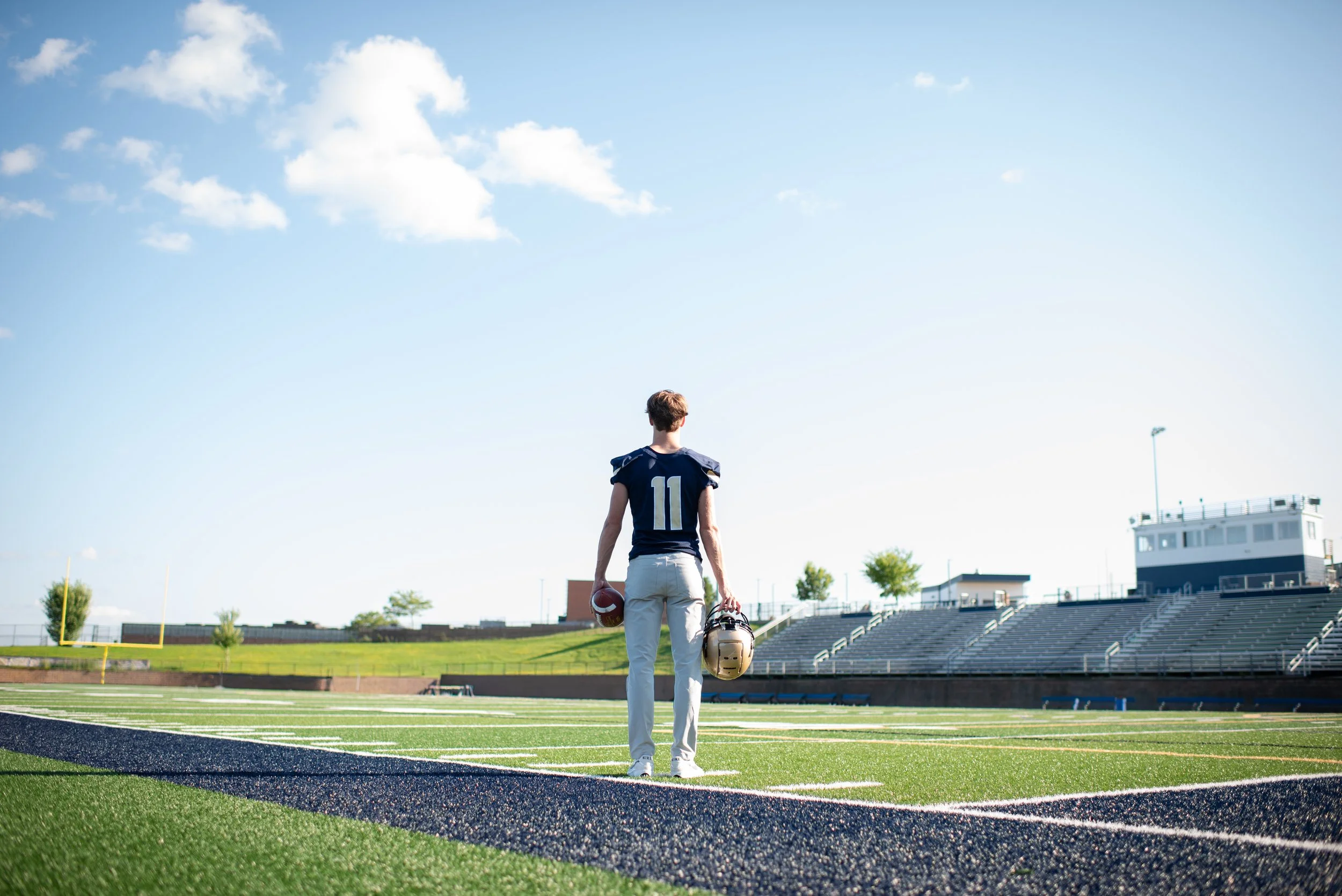 back side of senior boy staring at football field holding helmet and football