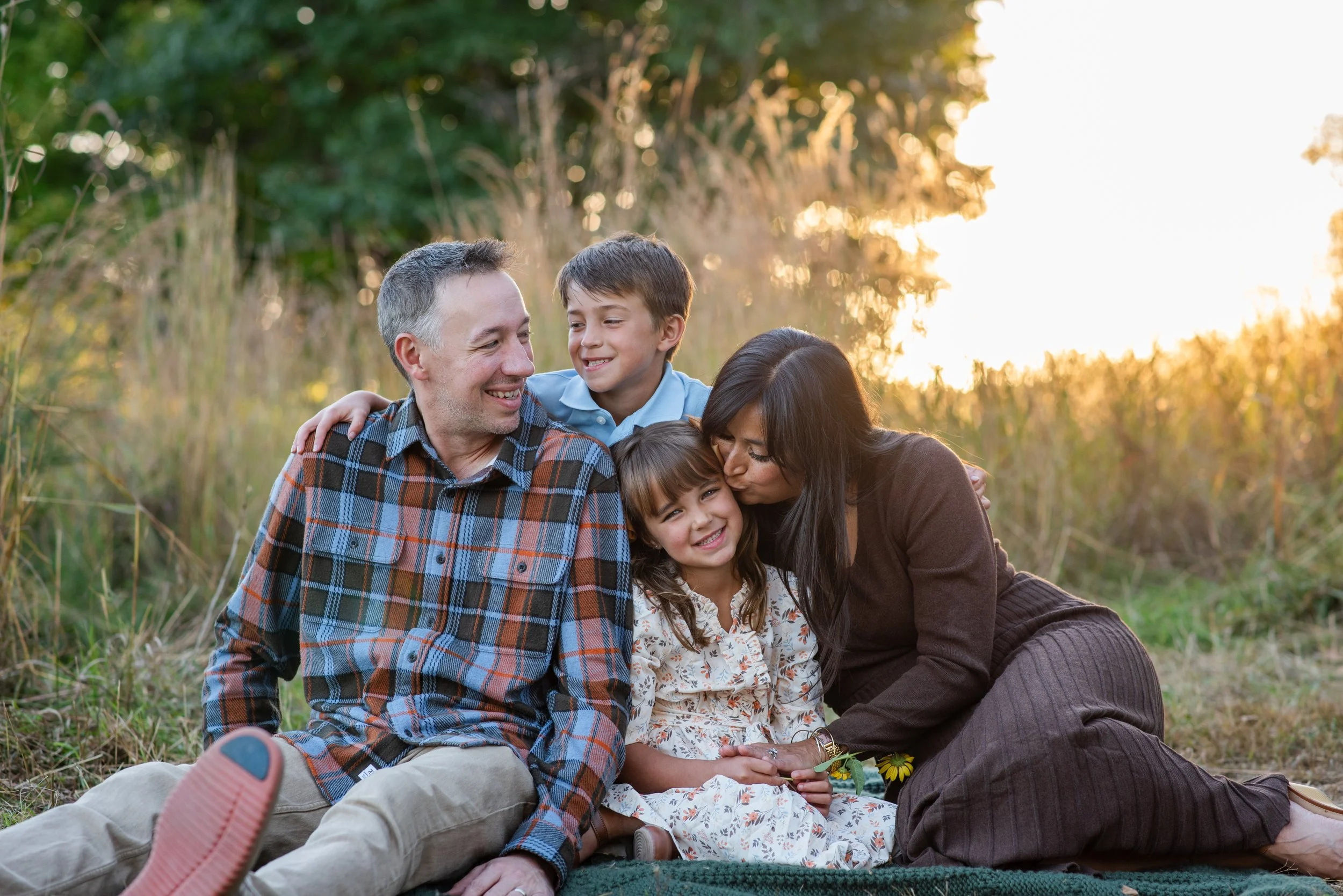 family of four sitting in front of tall grass with sun setting behind them