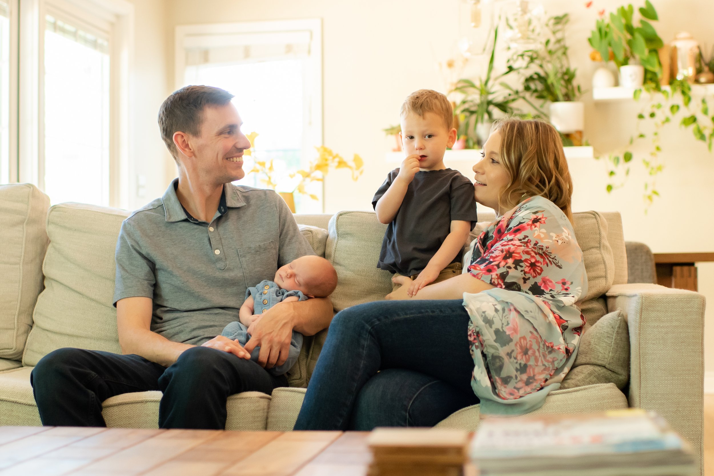 family cuddled up on couch while baby sleeps and toddler smiles during newborn photo session