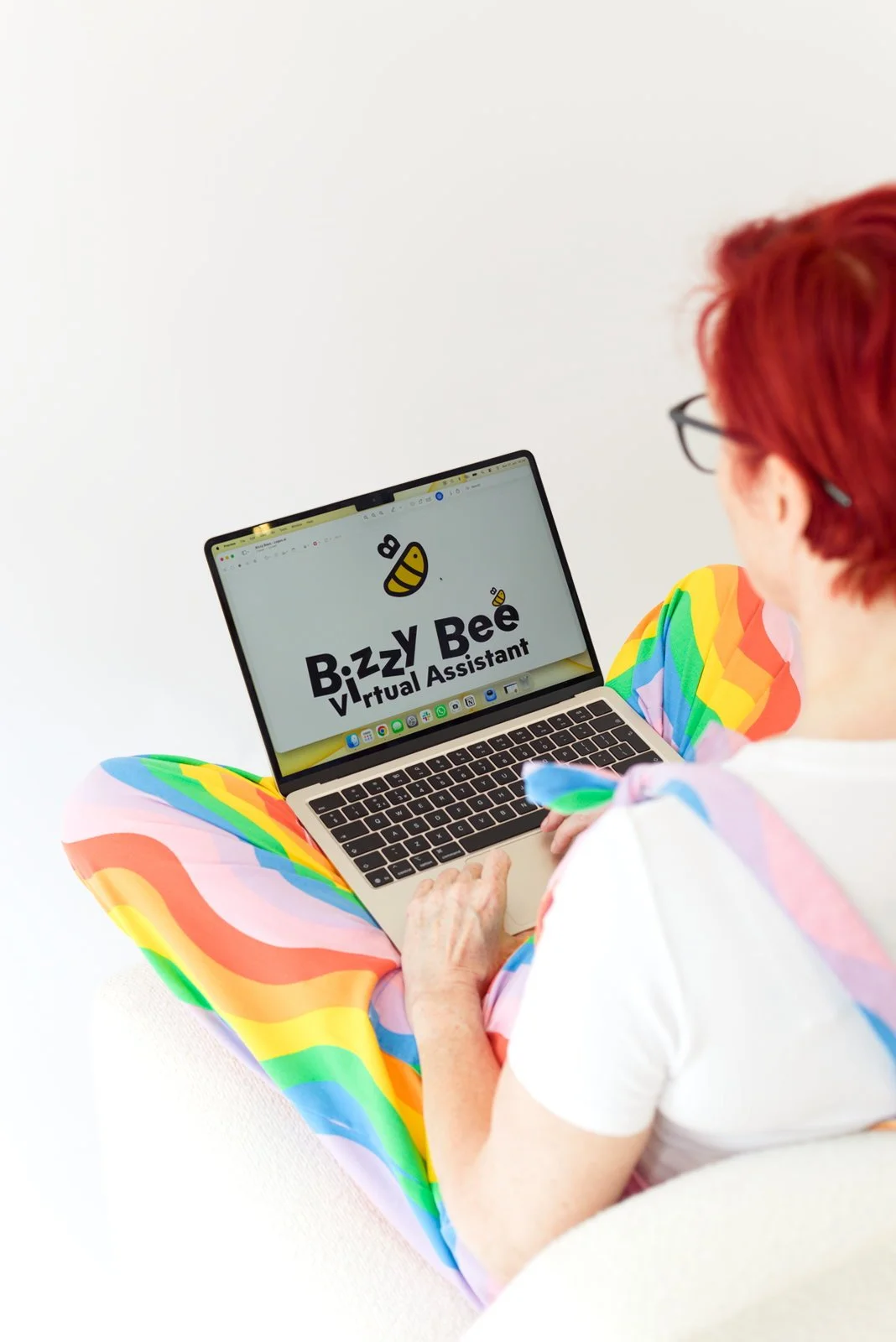 A person with red hair and glasses, wearing colorful rainbow pants and a white shirt, sitting cross-legged on the floor, looking at a laptop displaying the logo for "Buzzy Bee Virtual Assistant."