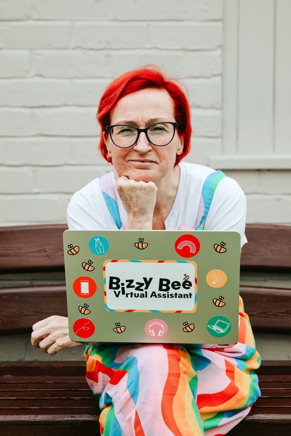 A person with red hair and glasses sitting on a wooden bench outdoors, holding a laptop labeled 'Bizzy Bee Virtual Assistant' decorated with bee and technology icons.