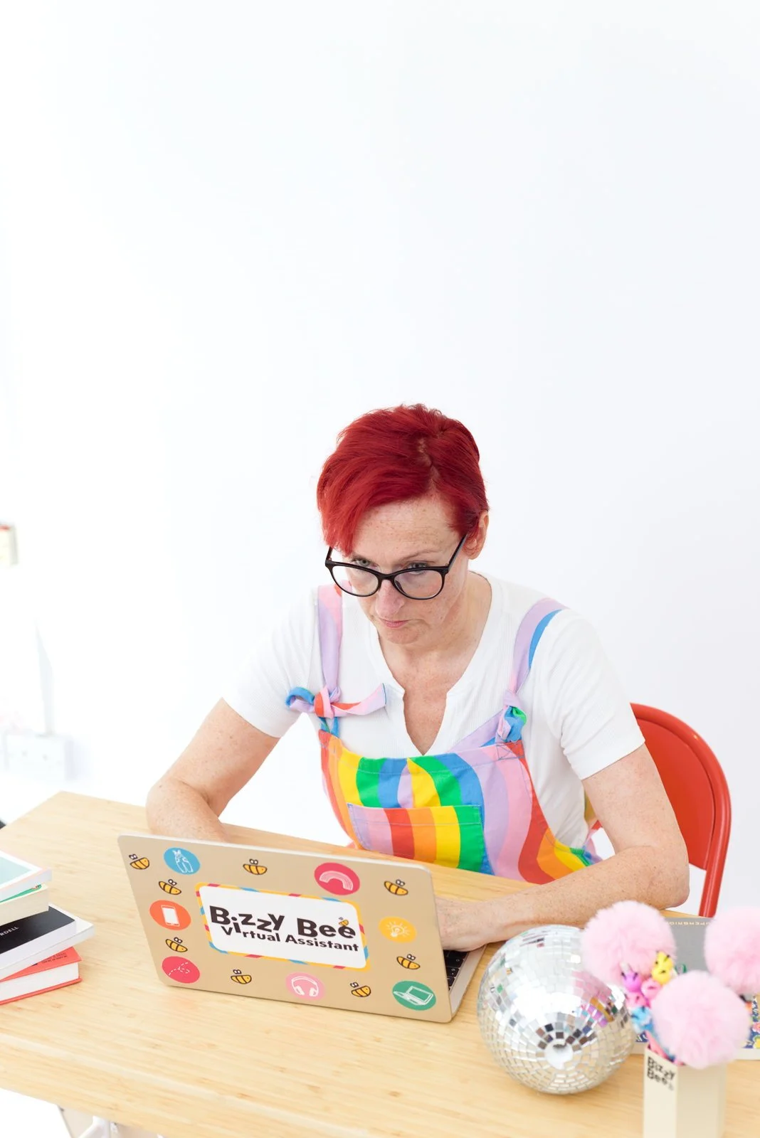Woman with red hair and glasses using a laptop at a desk, wearing a rainbow-colored apron, with colorful decorations and books nearby.