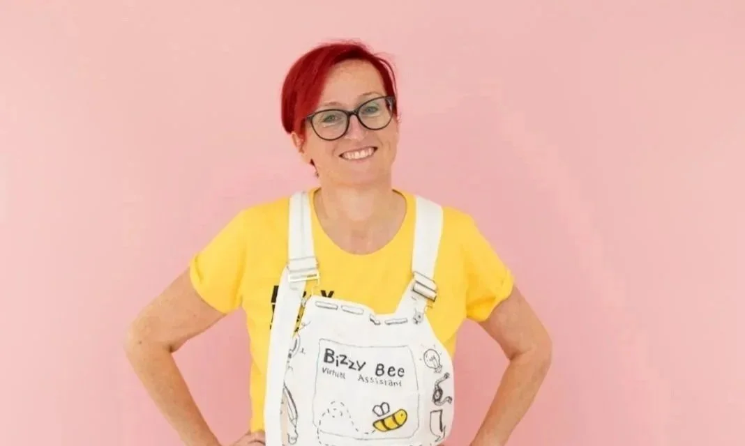 Woman with red hair, glasses, wearing a yellow T-shirt and white overalls with Bee illustrations, standing against a pink background, smiling.