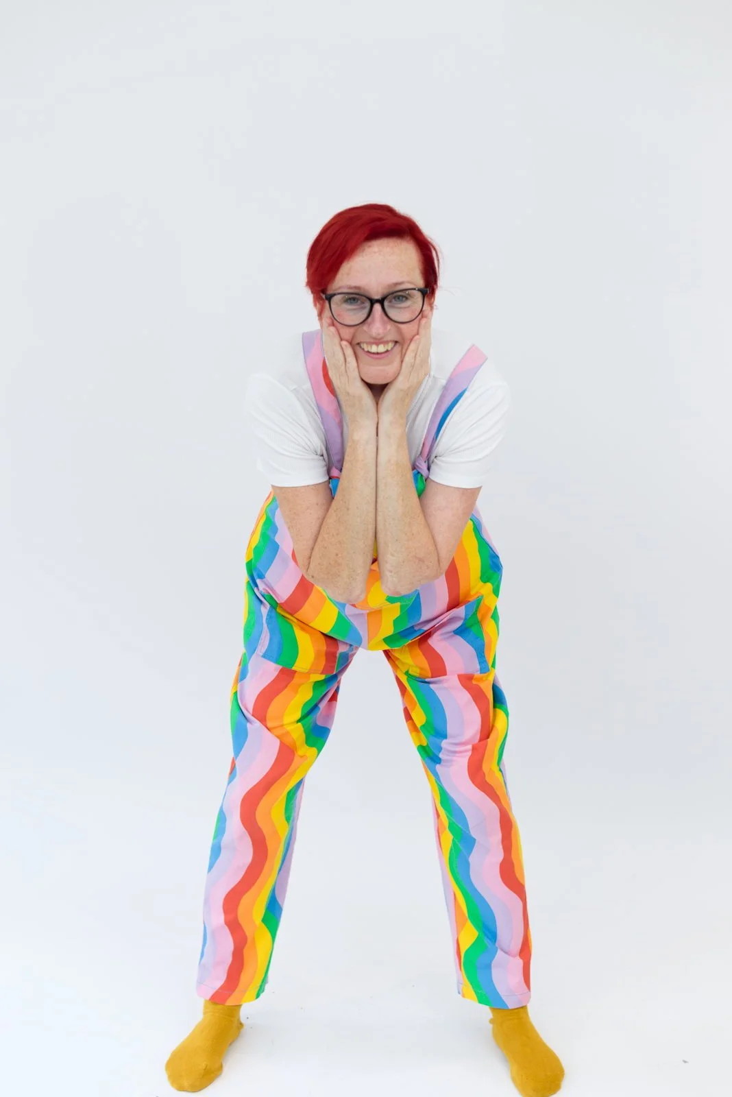 Person with red hair, glasses, wearing rainbow-striped pants and a white t-shirt, smiling and leaning forward with hands on cheeks against a plain white background.