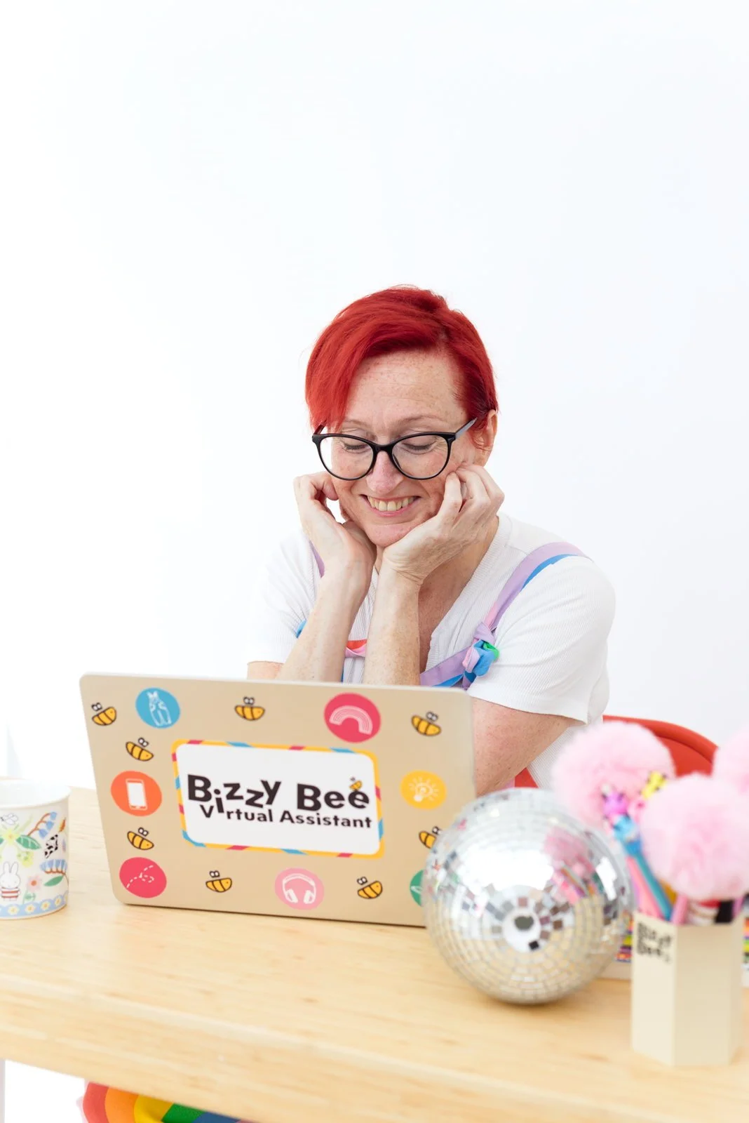 A woman with red hair, glasses, and a white shirt, smiling and holding her face as she looks at a colorful laptop with a sticker that reads 'Bizzy Bee Virtual Assistant.' The table has a mug with rainbow colors, a disco ball, and a container with pink fluffy pom-poms.