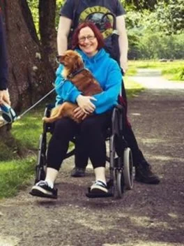 Woman sitting in a wheelchair holding a small brown dog outdoors in a park with a person standing behind her.