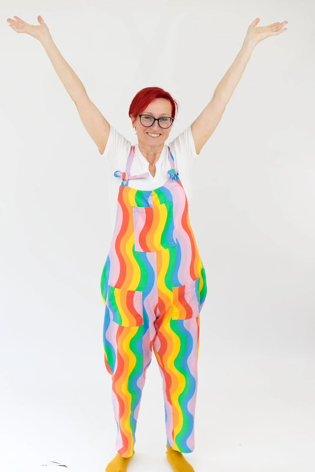 Woman with red hair and glasses wearing a rainbow-colored wavy pattern jumpsuit and yellow socks with arms raised in front of a white background.