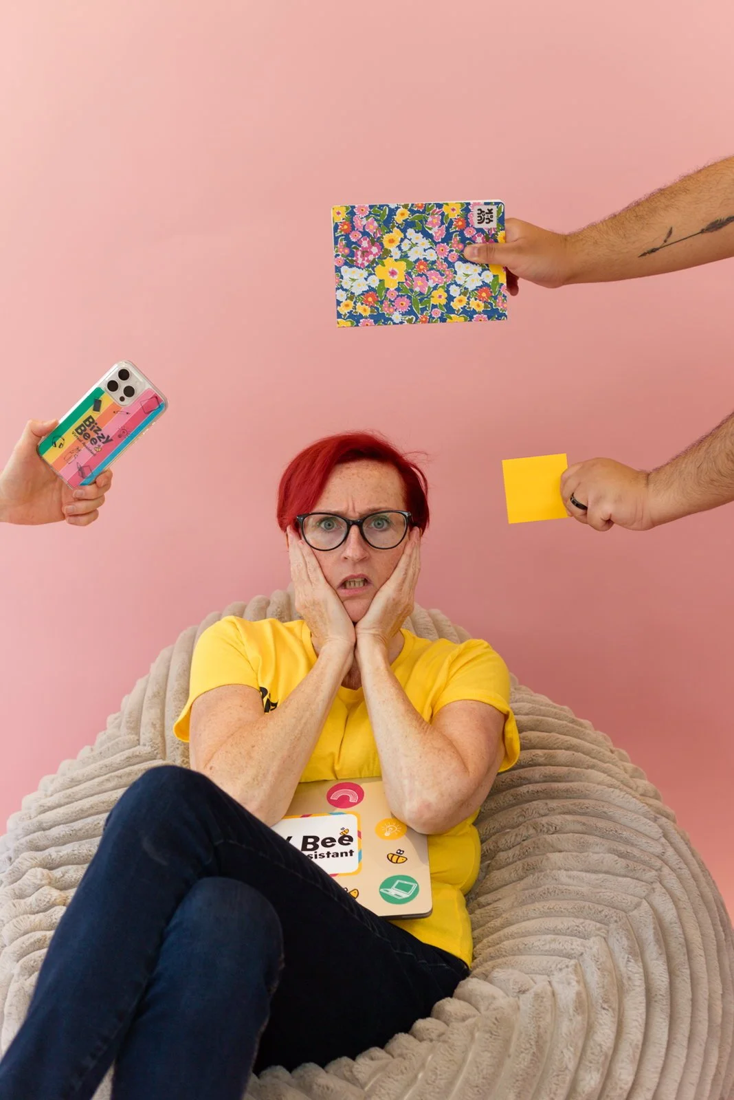 A woman with red hair and glasses sitting on a beige blanket, looking surprised or worried. Multiple hands are reaching toward her holding various objects, including a smartphone with a colorful case, a floral notebook, a yellow sticky note, and a yellow card.
