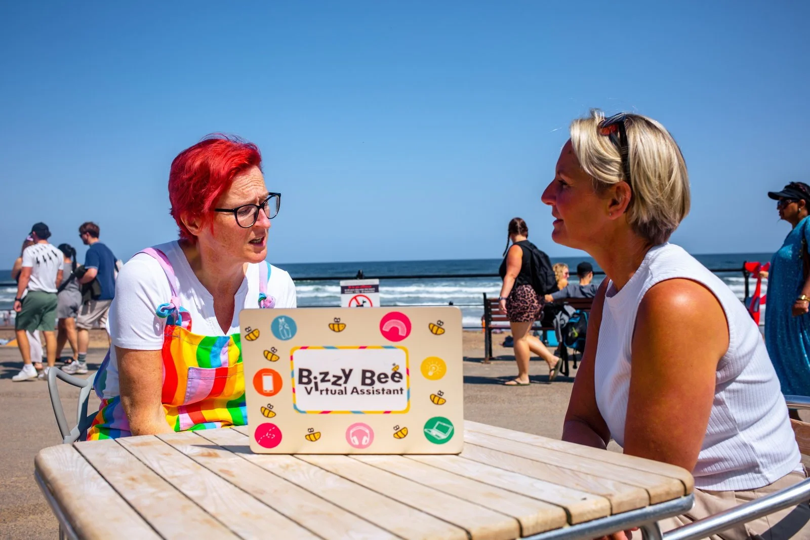 Two women are sitting at a table on a beach boardwalk, talking. One woman has short red hair and glasses, and wears a rainbow apron. The other woman has short blond hair, sunglasses on her head, and a white sleeveless top. There are beachgoers and the ocean in the background.