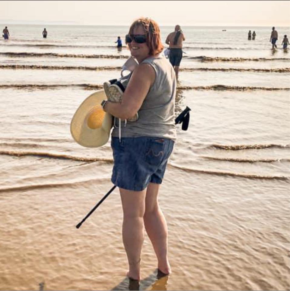 A woman with red hair, sunglasses, gray tank top, and blue shorts standing in shallow water at the beach, holding a large straw hat, goggles, and a cane, with people swimming and walking in the background.