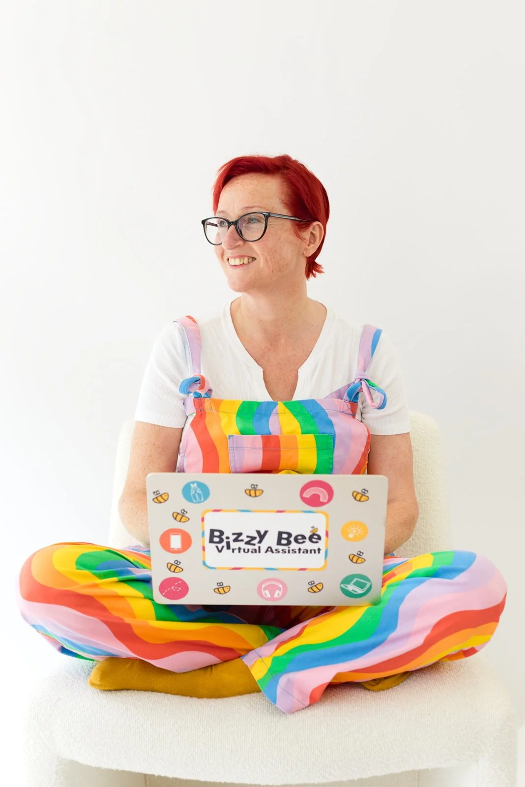 Woman with red hair and glasses sitting cross-legged on a white chair, wearing rainbow-colored pants and a rainbow apron, holding a laptop labeled 'Bizzy Bee Virtual Assistant' with various icons around it.