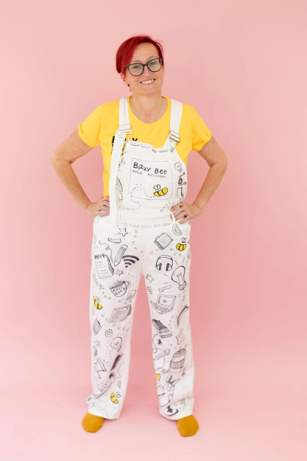A smiling woman with red hair, glasses, wearing yellow shirt and white overalls with doodles of electronic devices, books, and a bee, standing against a pink background.