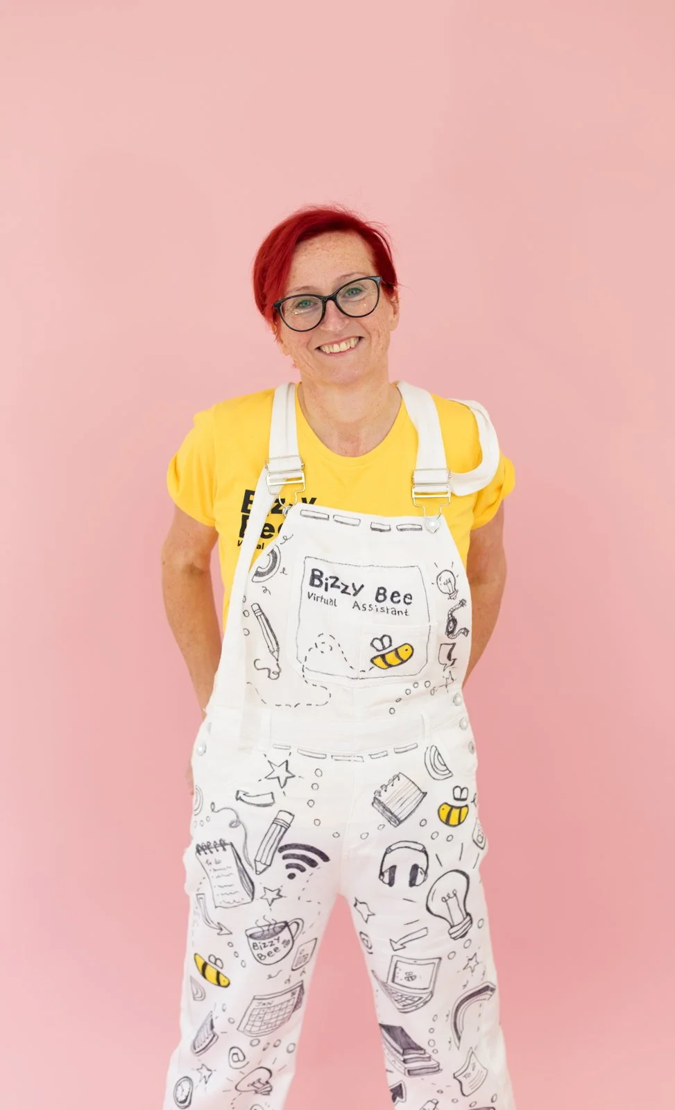 A woman with short red hair, glasses, and freckles, smiling against a pink background. She is wearing a yellow t-shirt and white overalls with black and yellow doodle sketches related to technology, creativity, and learning. The overalls have a patch on the front that reads "Bizzy Bee Virtual Assistant."