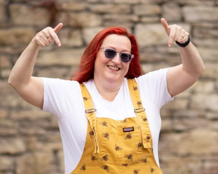 Smiling woman with red hair wearing sunglasses and bright yellow dungarees with bee prints, pointing downward with both hands while standing in front of a stone wall.