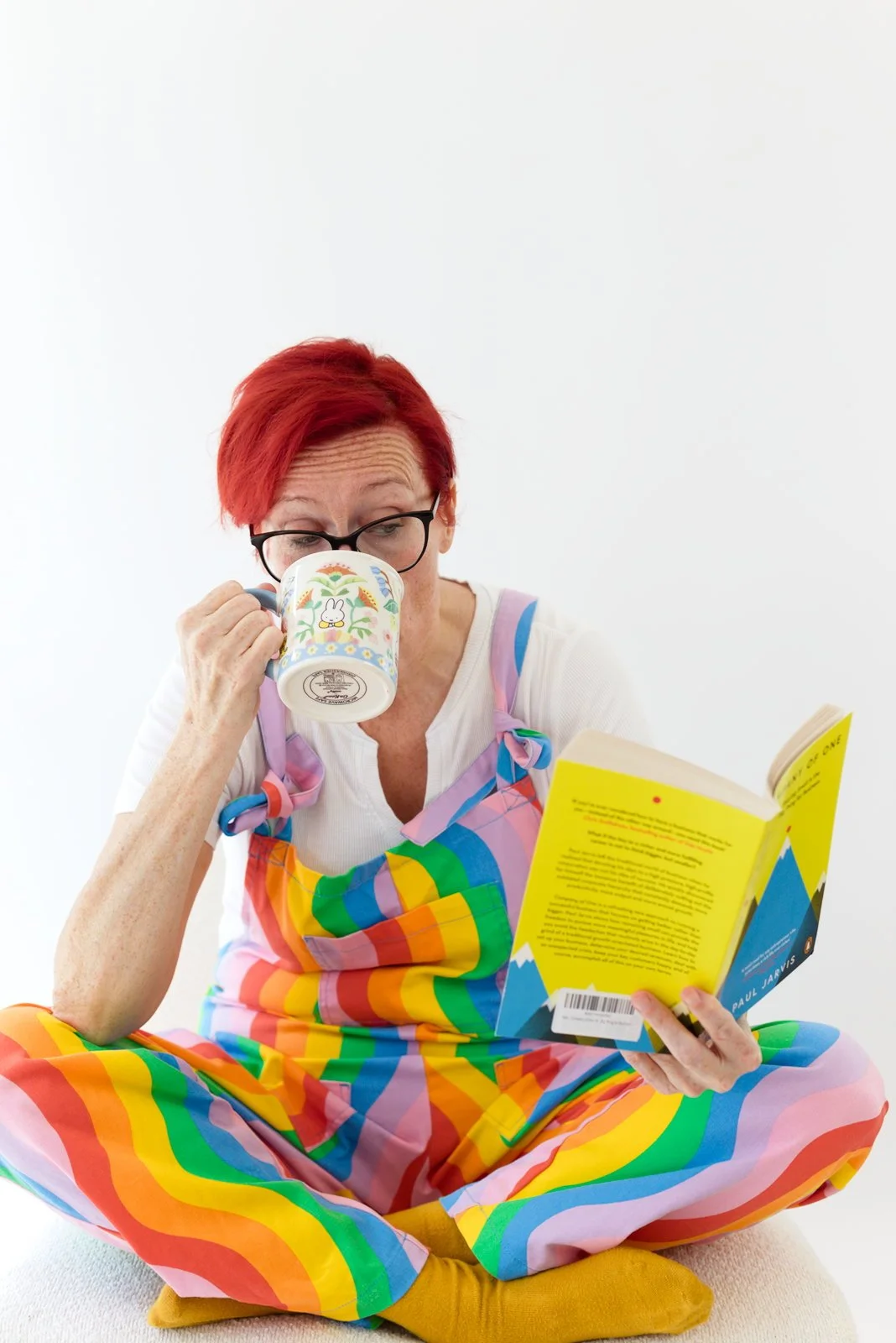 Woman with red hair and glasses sitting cross-legged, wearing rainbow-colored overalls, drinking from a mug with a bunny design, and reading a book.