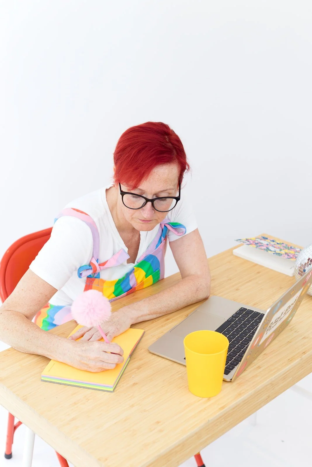 A woman with red hair, glasses, and freckles, wearing a colorful rainbow apron, sits at a wooden desk with a laptop, yellow cup, notebook, and snacks, writing with a pink fluffy pen.