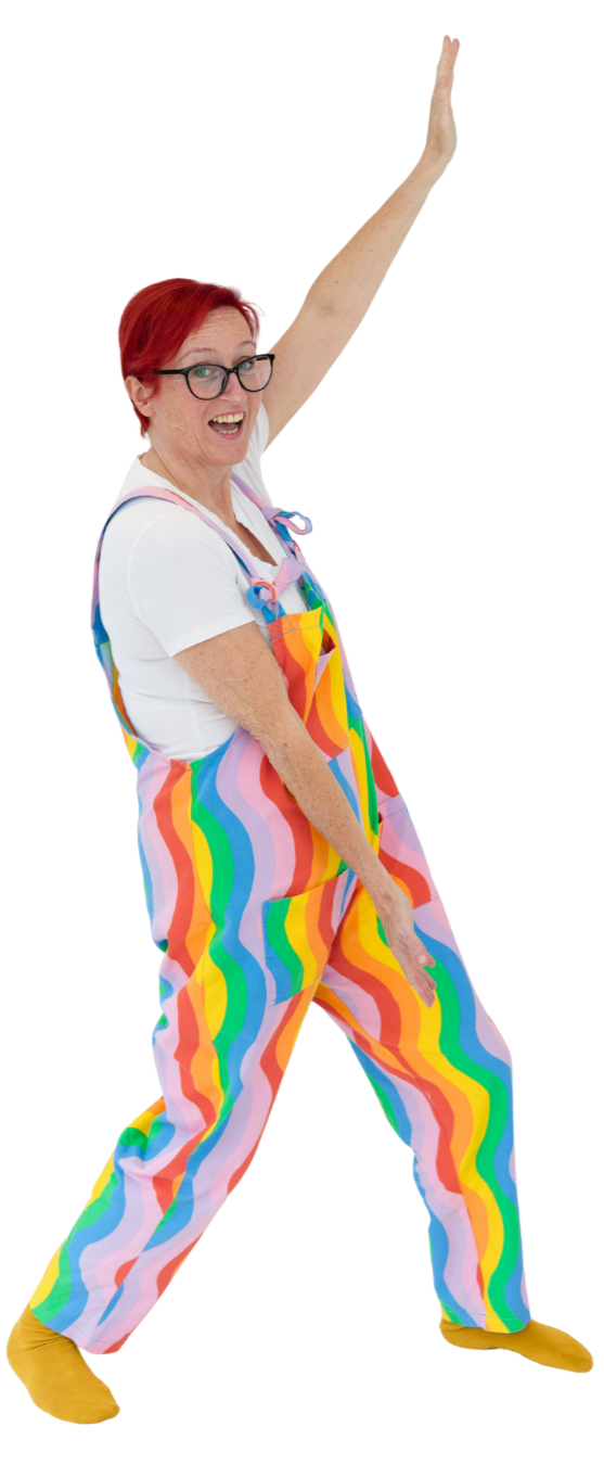 A woman with red hair, glasses, and a big smile, wearing colorful rainbow-striped overalls, a white shirt, yellow slippers, and posing with one arm raised and the other pointing down.