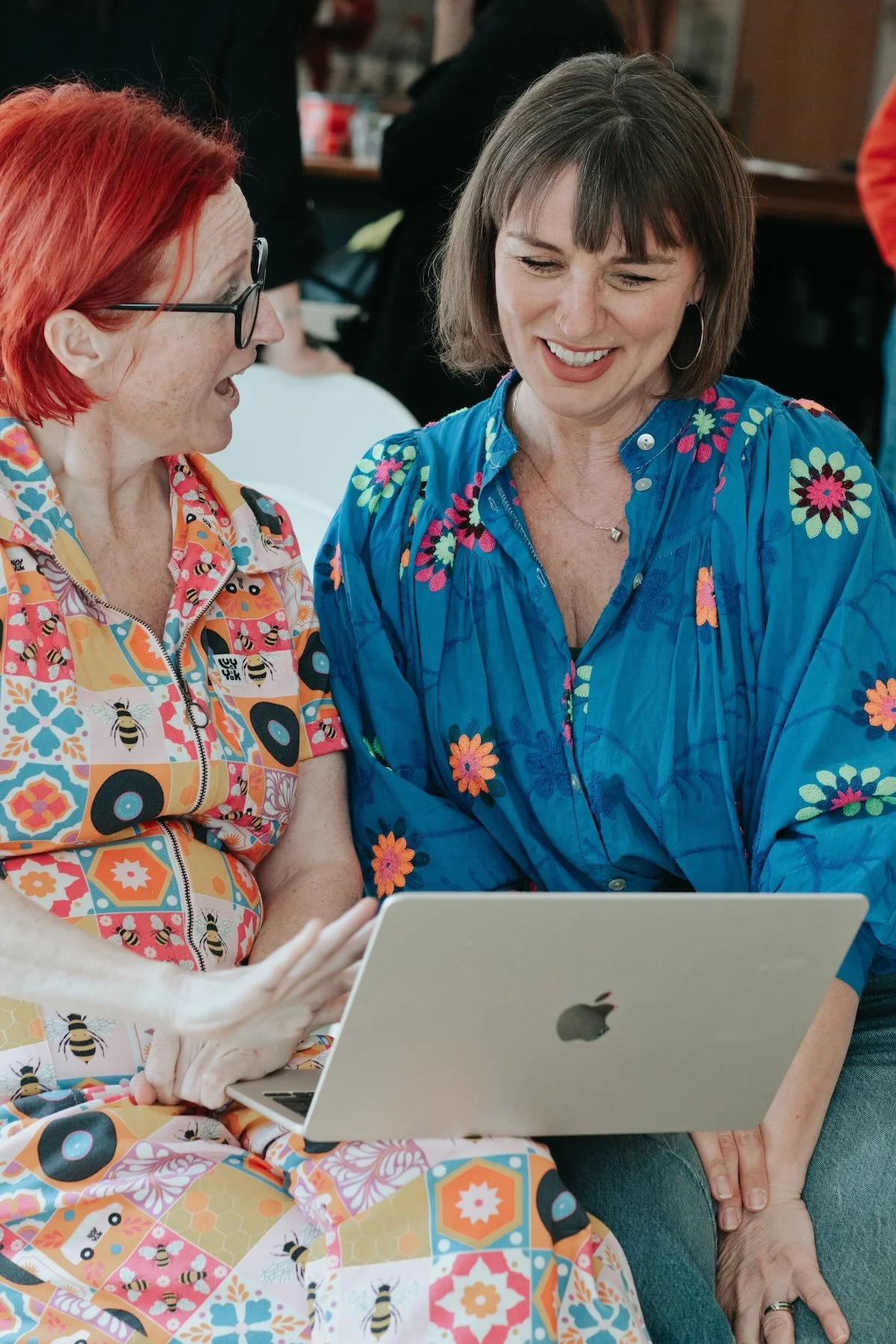 Two women sitting closely, looking at a silver MacBook laptop, smiling and engaging with each other, in a social setting.