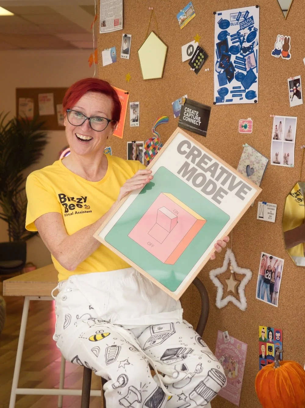 A woman with red hair, glasses, and a bright yellow shirt labeled 'Bizzy Bee Virtual Assistant' is smiling and holding a sign that reads 'CREATIVE MODE' with an illustration of an on-off switch. She is sitting on a stool in front of a corkboard decorated with photos, colorful paper cutouts, and posters, including one saying 'CREATE CAPTURE CONNECT'.
