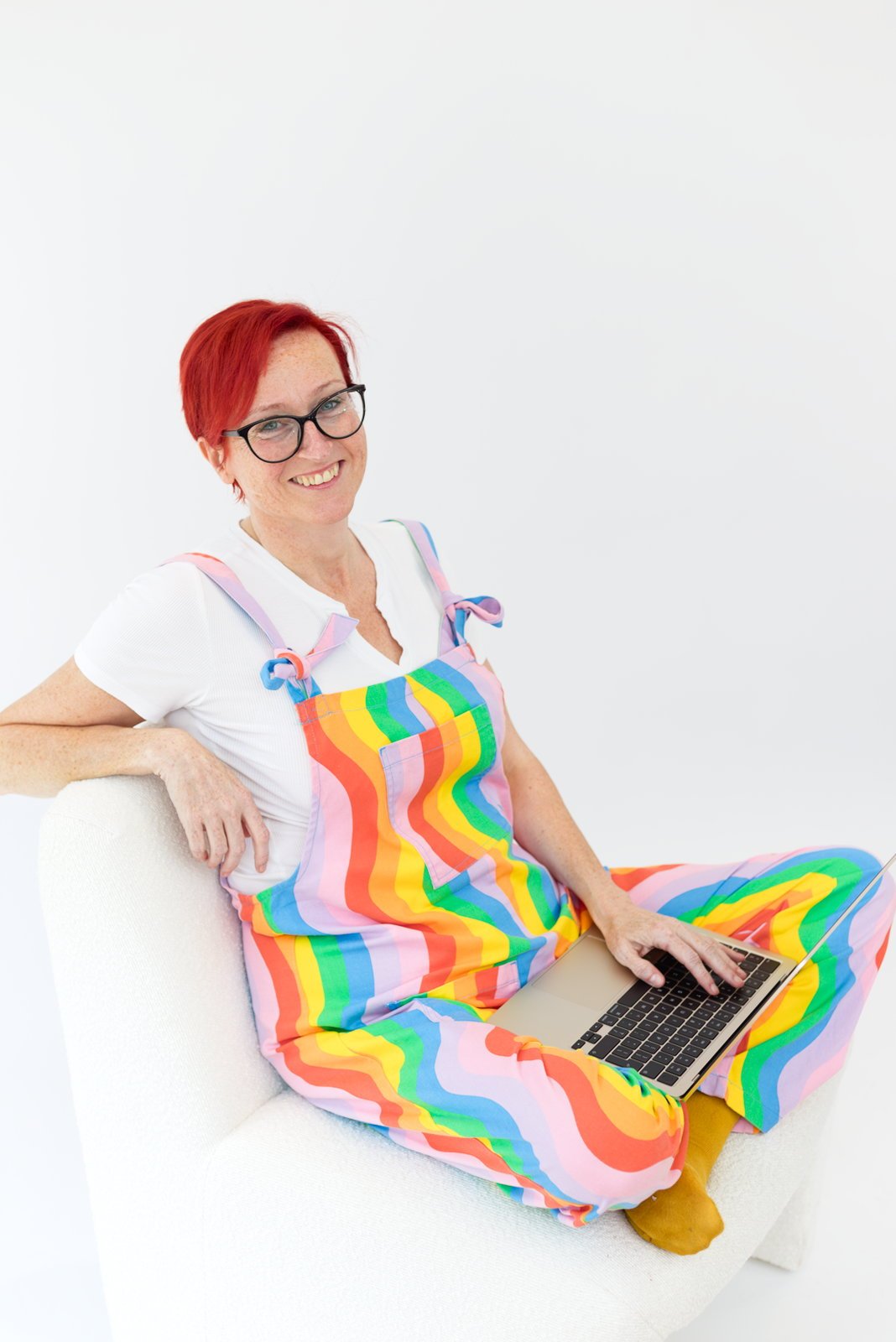 Woman with red hair and glasses sitting on a white couch, wearing a rainbow-striped overall, white shirt, and yellow socks, smiling while using a laptop.