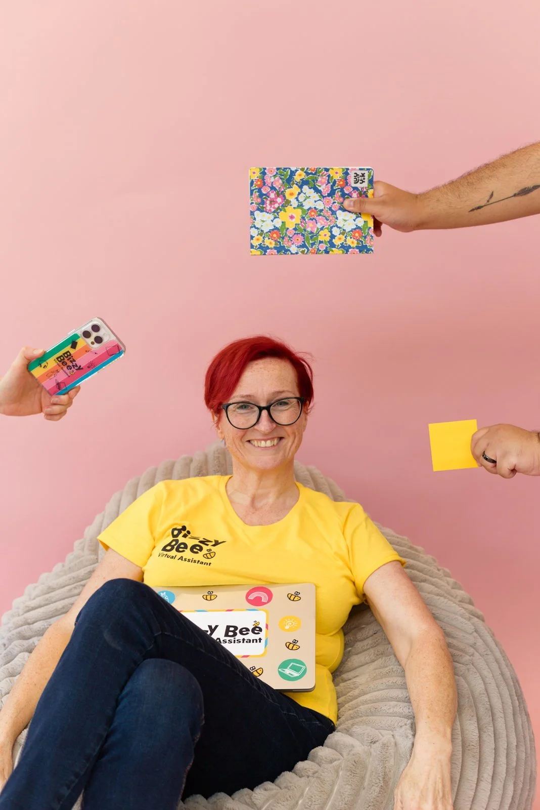 Erin Buck smiling on a bean bag with a BizzyBee Virtual Assistant tablet, surrounded by hands holding notes and a phone