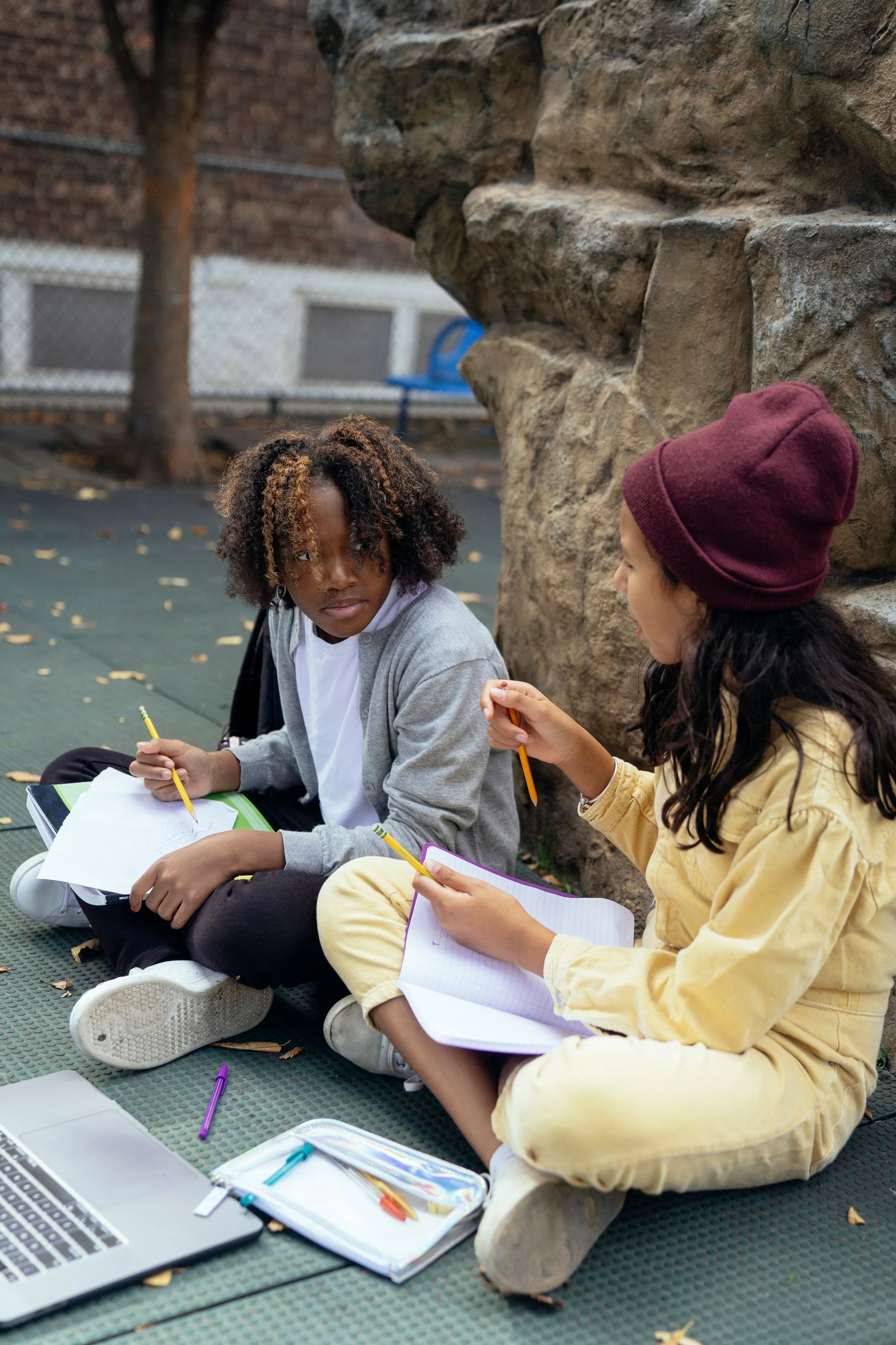 Two children sitting on the ground outside, studying with notebooks and pencils, near a stone wall and a tree, on a cold day.