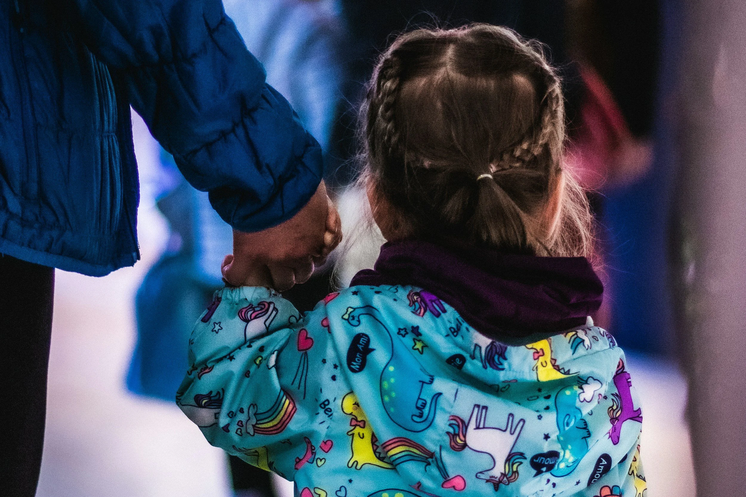 A young girl with braided hair wearing a colorful jacket with unicorn and rainbow patterns, being held or supported by an adult in a blue jacket.