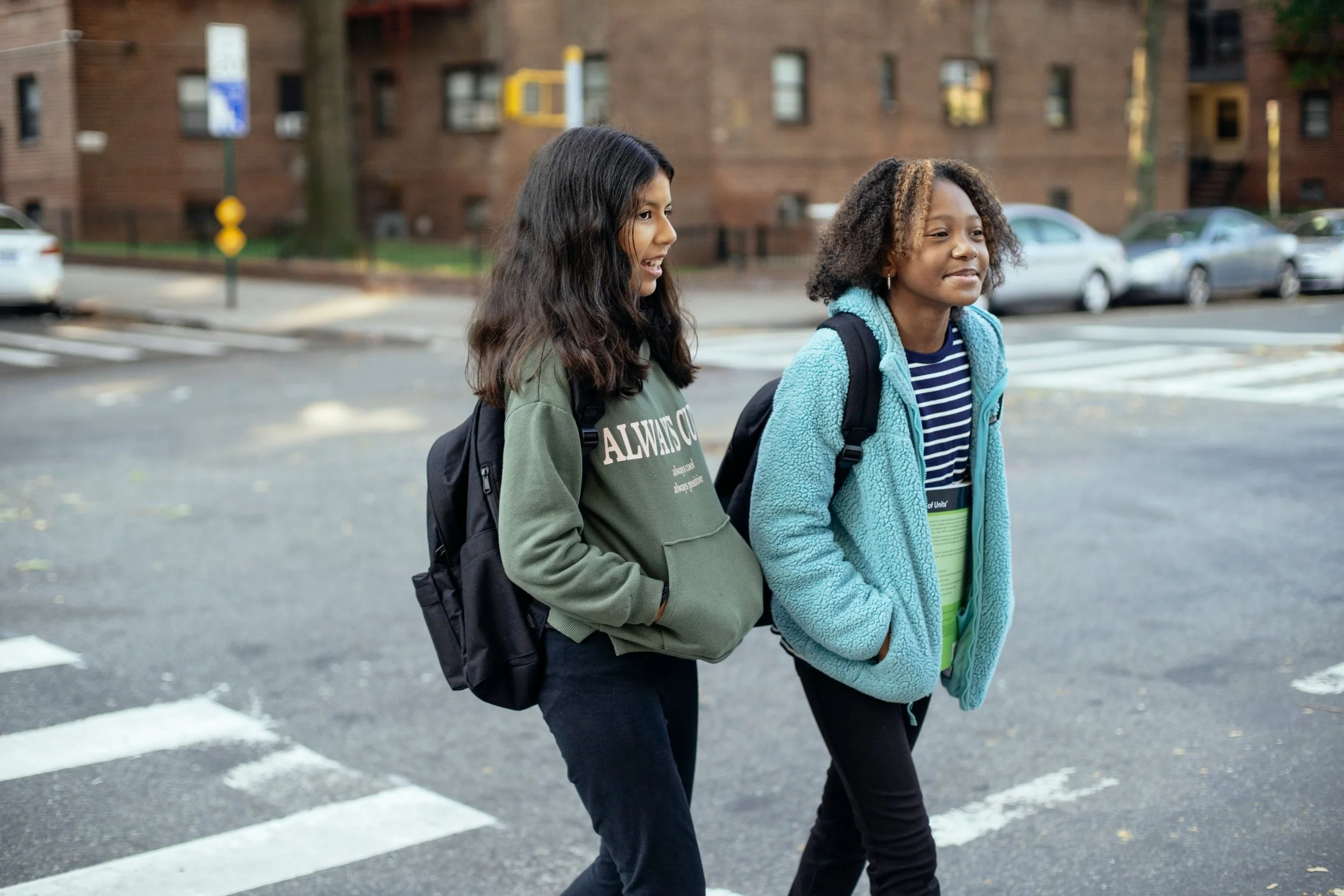Two young girls walking across a crosswalk in an urban neighborhood, wearing casual clothing and backpacks, with brown brick apartment buildings in the background.