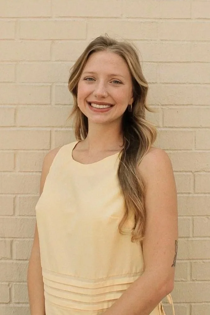 Young woman with long wavy hair, smiling, wearing a sleeveless light yellow dress, standing in front of a beige brick wall.