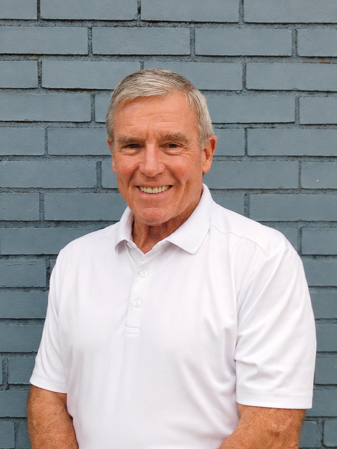 Garner Tullis, the founder of Our Greater Good. An older man with gray hair, smiling, wearing a white polo shirt, standing in front of a gray brick wall.