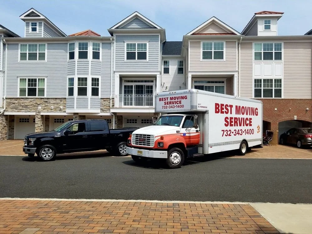 Moving truck labeled 'Best Moving Service' parked on the street in front of a modern apartment complex with multi-story buildings and a brick facade.