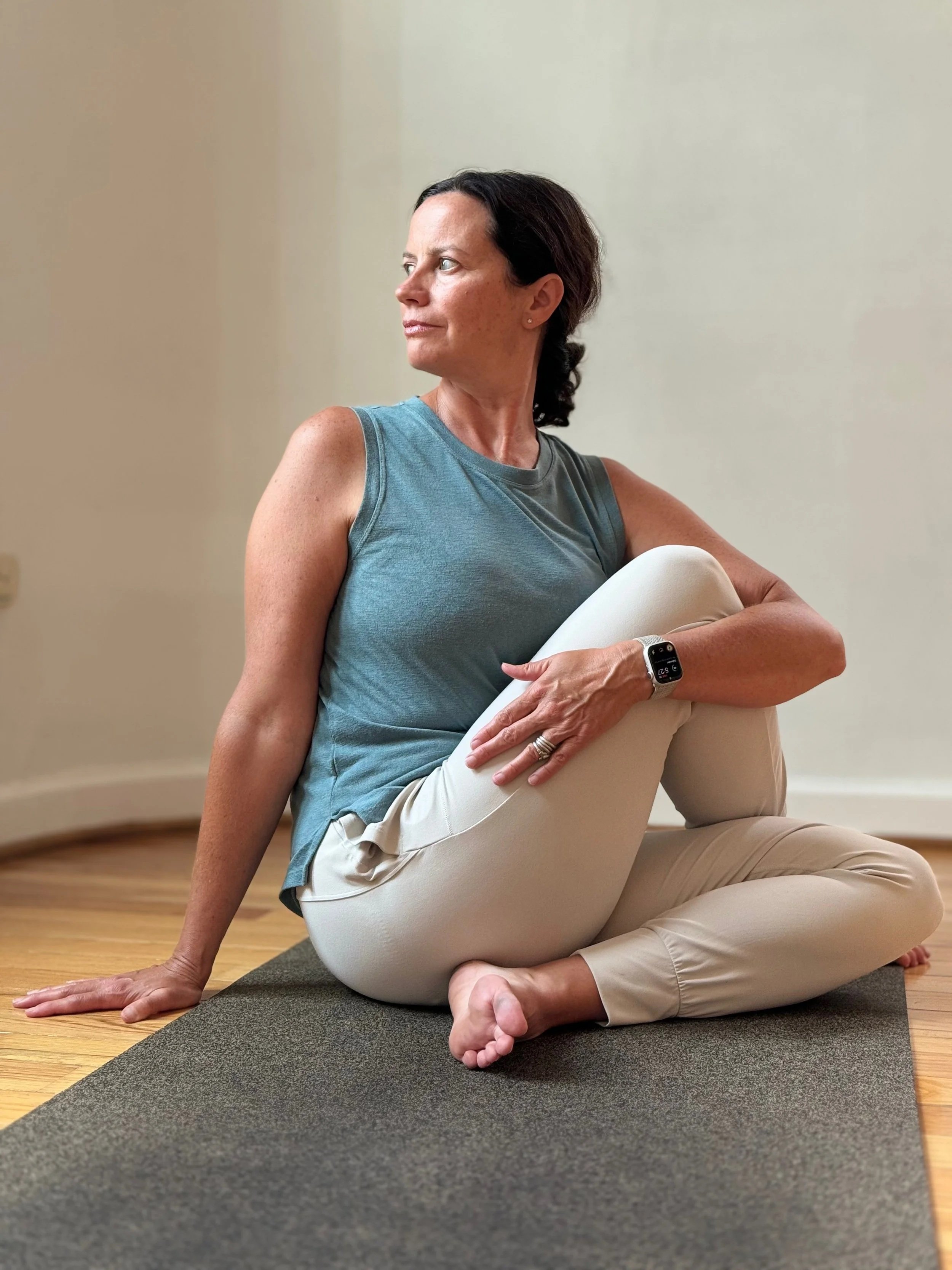 A woman practicing yoga indoors on a black mat, sitting cross-legged with one hand on the floor and the other arm wrapped around her bent knee, looking to the side.
