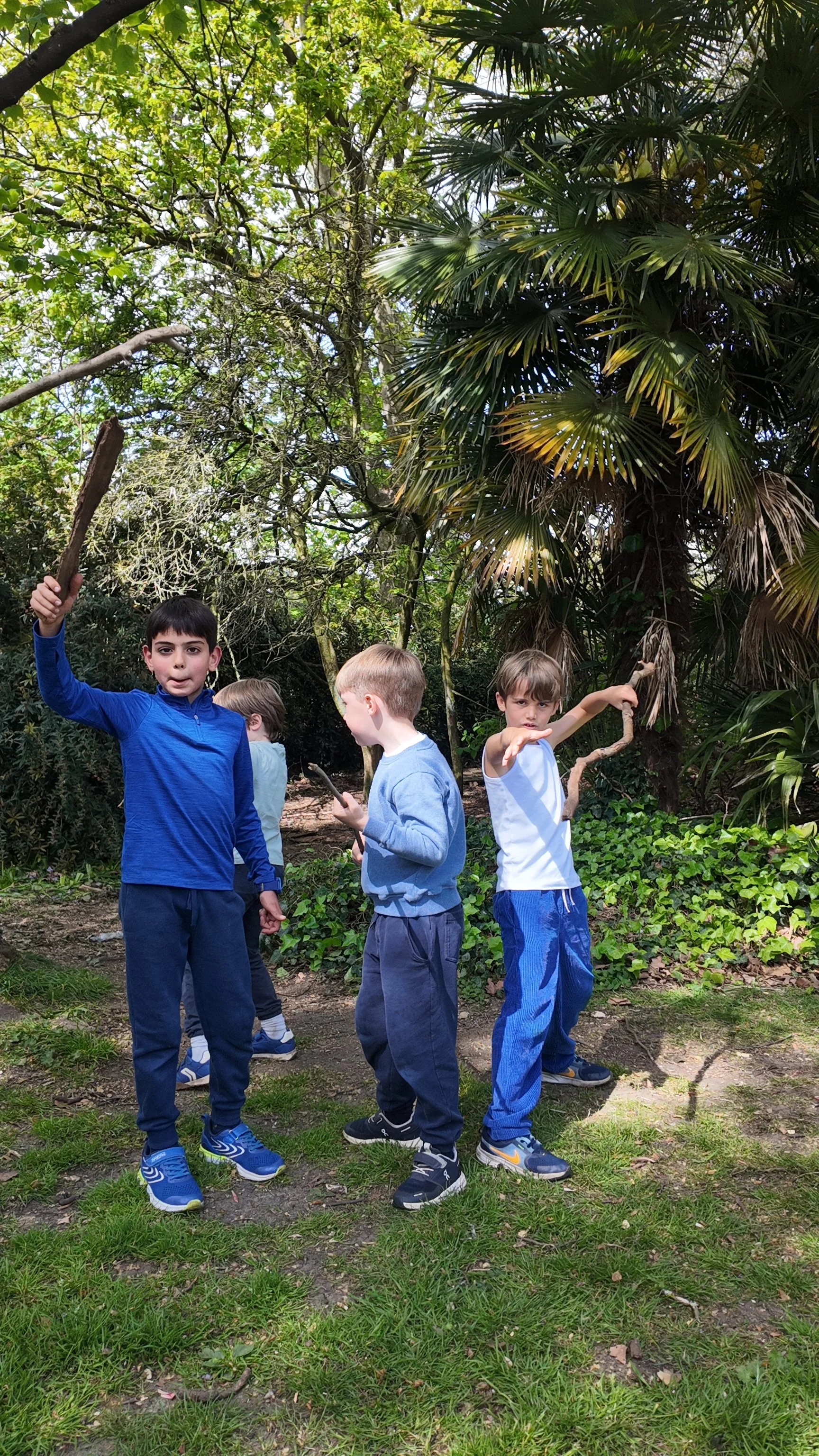 Four young boys standing in a lush, green outdoor setting with trees and plants, some holding sticks or branches.