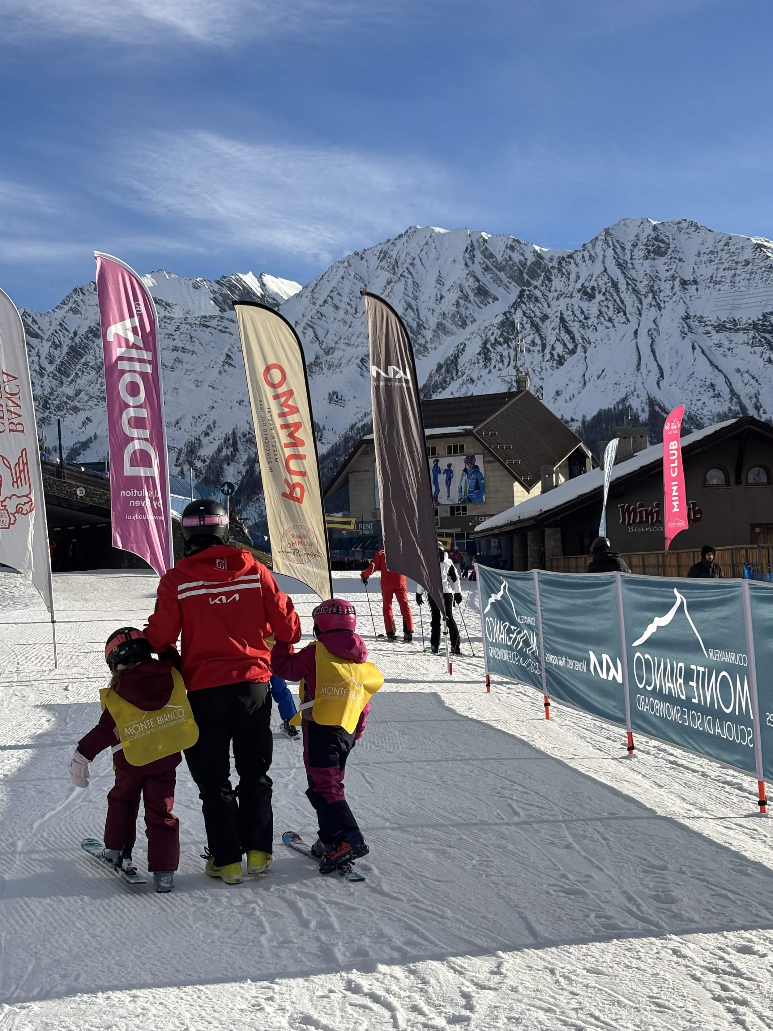 Children on skis learning with an instructor at a ski resort with snow-covered mountains in the background, colorful flags, and a building, happy children during the kids club with Archies family ski week in Courmayeur