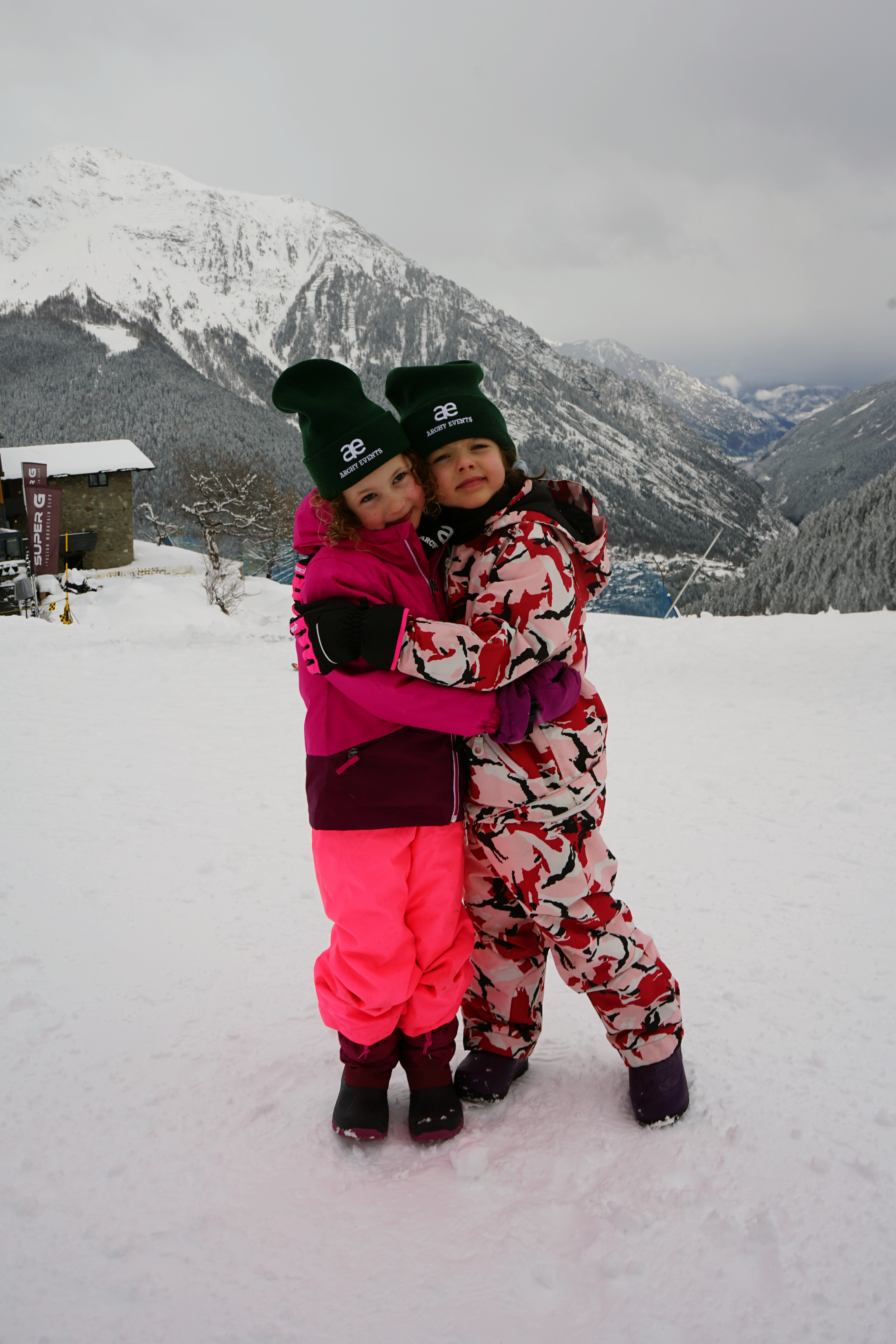 Two children in colorful winter outfits hugging on a snowy mountain landscape with snow-covered mountains and cloudy sky in the background.