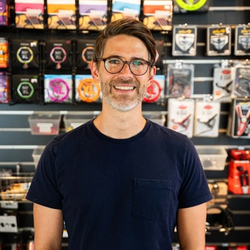 A man with glasses and a beard smiling in a sporting goods store, with various bicycle accessories hanging on the wall behind him.
