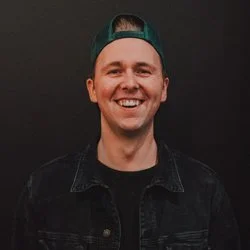 A young man smiling, wearing a black jacket and a backward green cap, against a dark background.