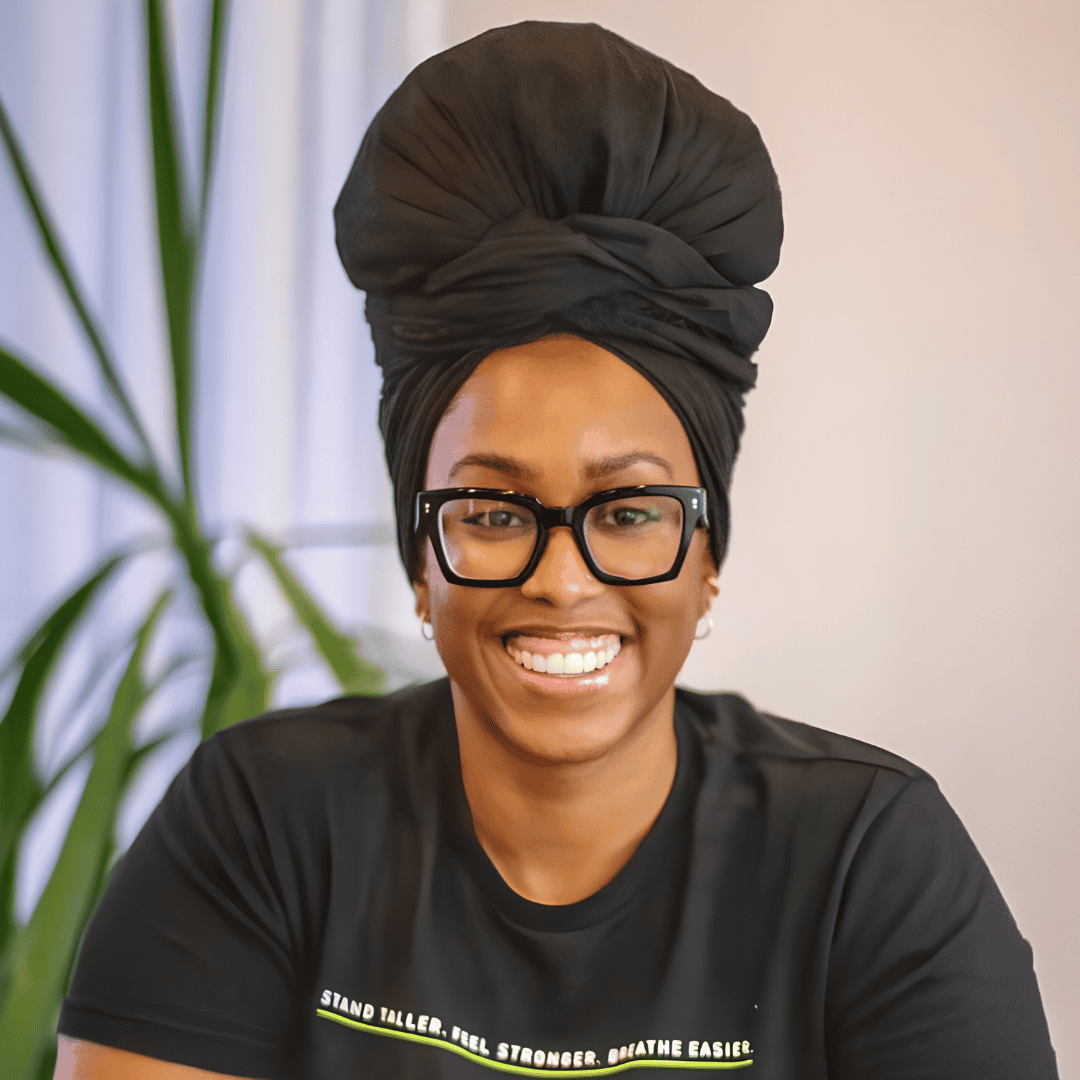 A smiling woman wearing black glasses and a black head wrap sits in a black t-shirt with a motivational slogan, with a plant in the background | ReEvolve Therapy