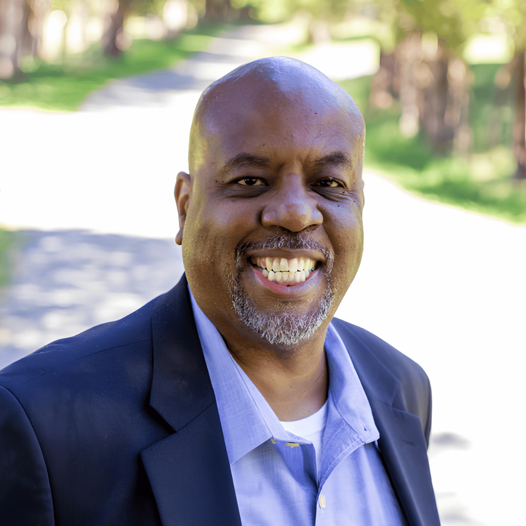 A smiling bald man with a gray beard wears a navy blazer over a light blue shirt, standing outdoors on a tree-lined path | ReEvolve Therapy