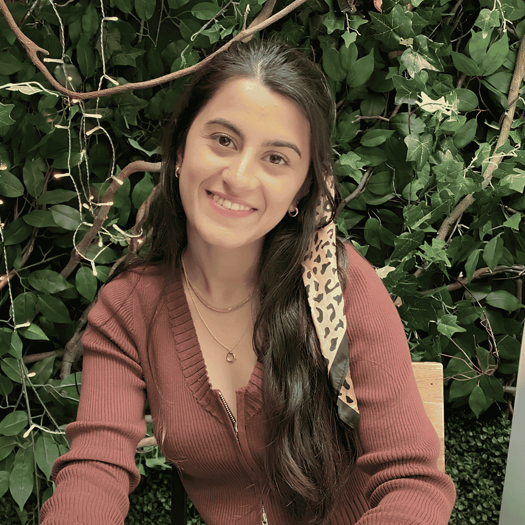 A smiling woman with long dark hair wears a rust colored sweater and layered necklaces, seated in front of a green foliage wall | ReEvolve Therapy