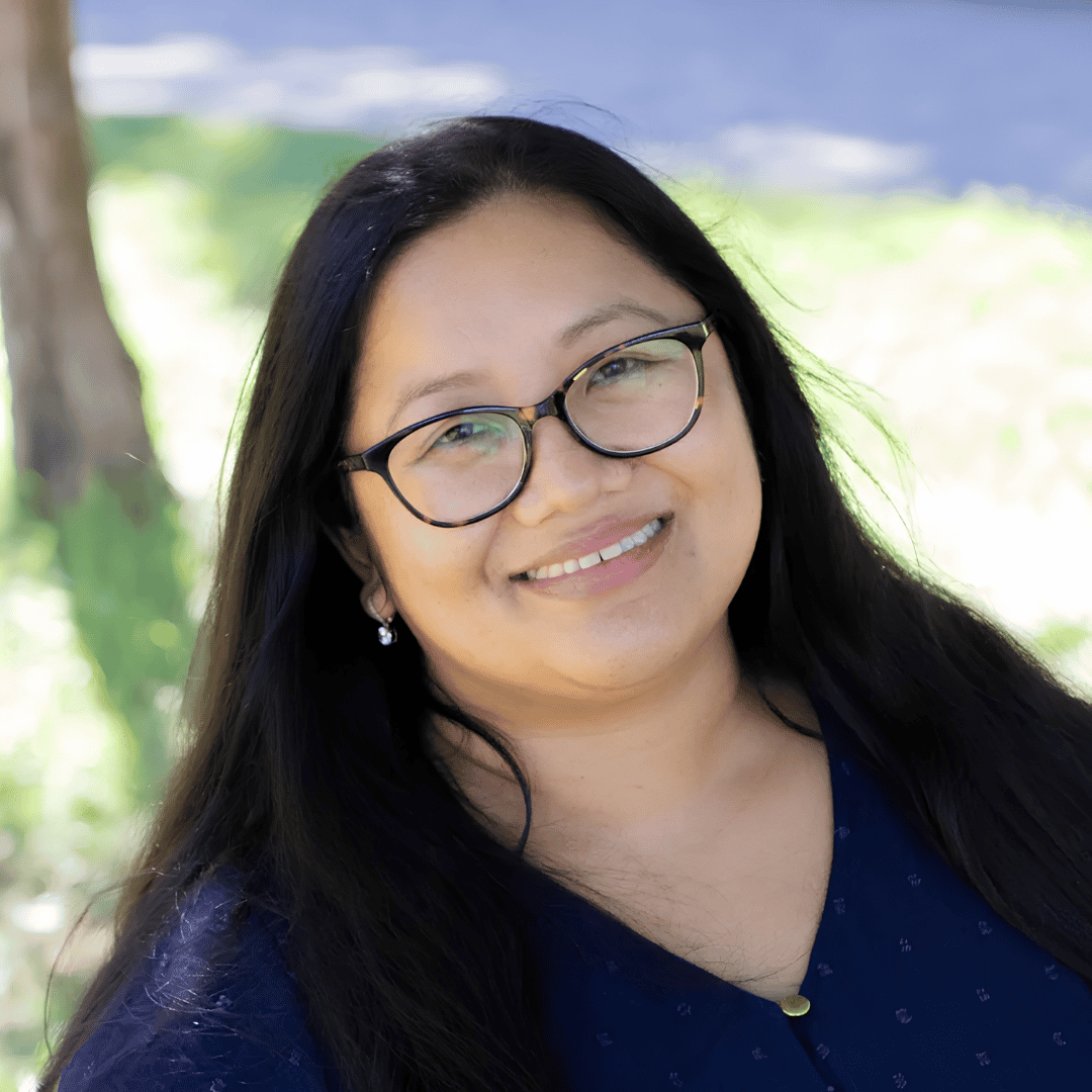A smiling woman with long dark hair wears tortoiseshell glasses and a navy patterned top, photographed outdoors near a tree | ReEvolve Therapy