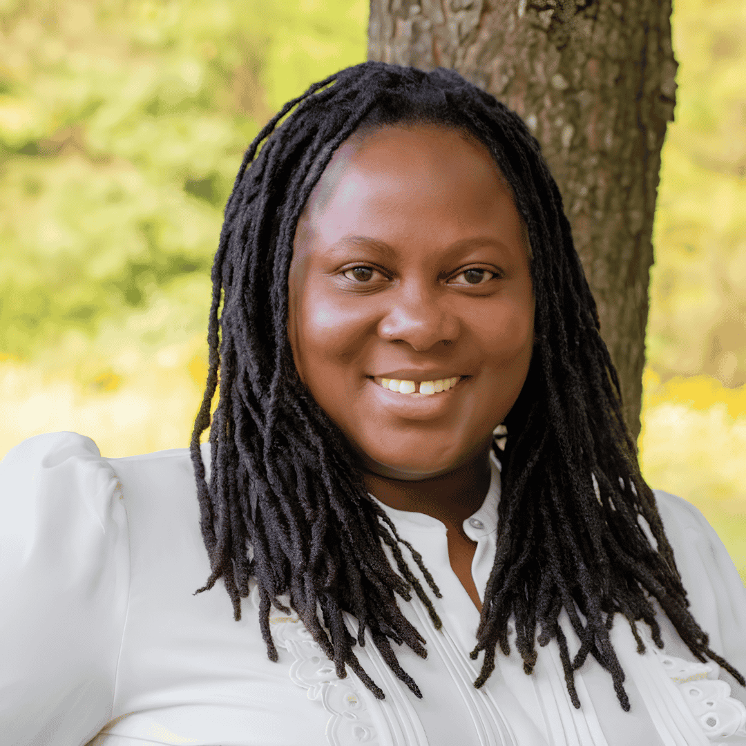 A smiling woman with long dreadlocks wears a white blouse with lace detailing, standing outdoors in front of a tree | ReEvolve Therapy