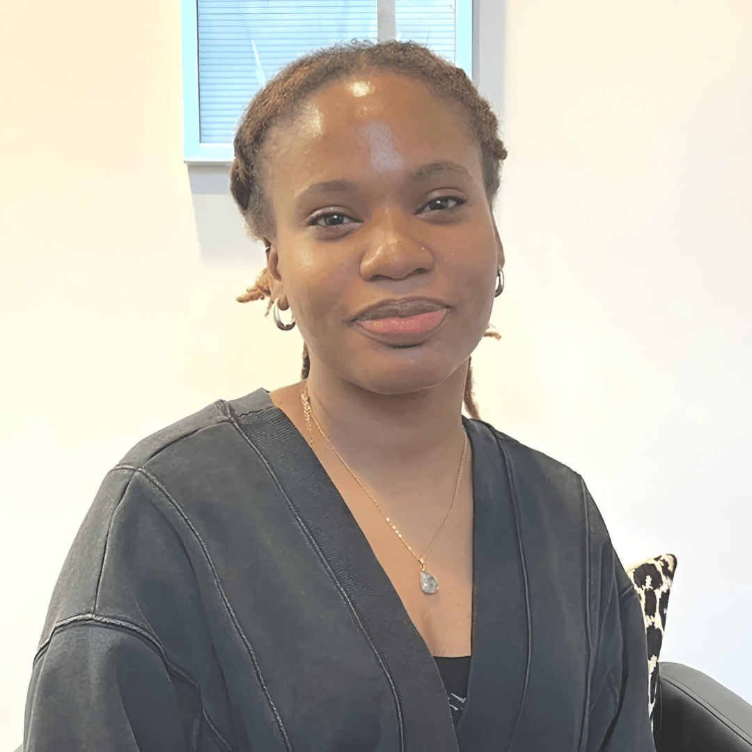 A smiling woman with braided hair wears a dark cardigan and a pendant necklace, seated indoors in a black chair | ReEvolve Therapy