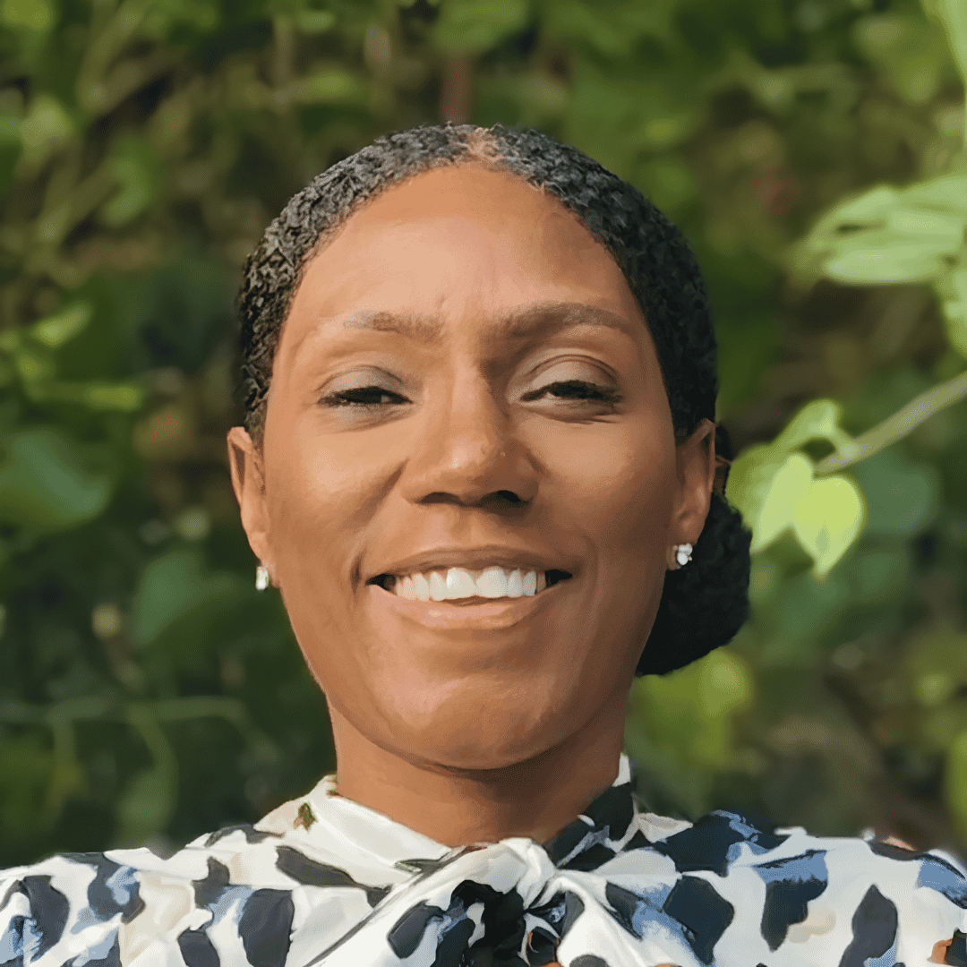 A smiling woman with hair pulled back wears a patterned blouse with a tied neckline, photographed outdoors with greenery in the background | ReEvolve Therapy